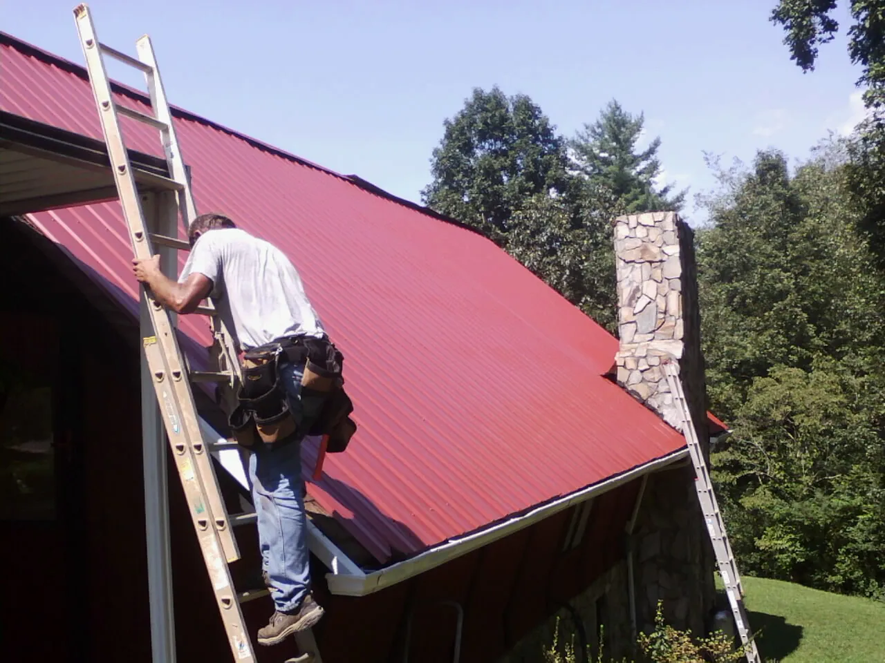 Roofer on a ladder beside a red metal roof and stone chimney — Soffit & Fascia Repair by Roofing Brothers in Canistota